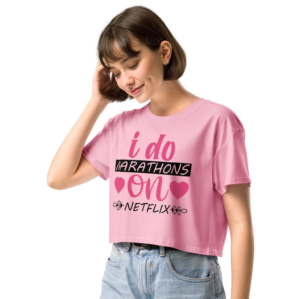 Ein "Bubblegum-Pink" farbenes, kurz geschnittenes Damen-T-Shirt (Crop Top) mit dem Text "i do MARATHONS on NETFLIX" auf der Vorderseite. Der Text "i do" und "on" ist in Pink, "MARATHONS" in Schwarz auf einem schwarzen Balken, und "NETFLIX" ebenfalls in Schwarz, umgeben von kleinen schwarzen Herzen und floralen Ranken.