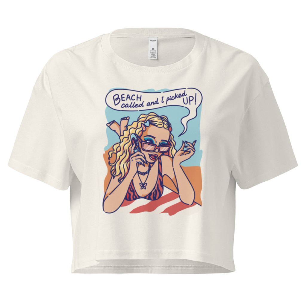 Ein ecru farbenes kurz geschnittenes Damen-T-Shirt (Crop Top) mit einer Retro-Illustration einer blonden Frau mit Sonnenbrille und gestreiftem Bikini, die entspannt am Strand liegt und telefoniert. Über ihr ist eine Sprechblase mit dem Text "BEACH called and I picked UP!" zu sehen. Der Hintergrund zeigt Sand und Meer.