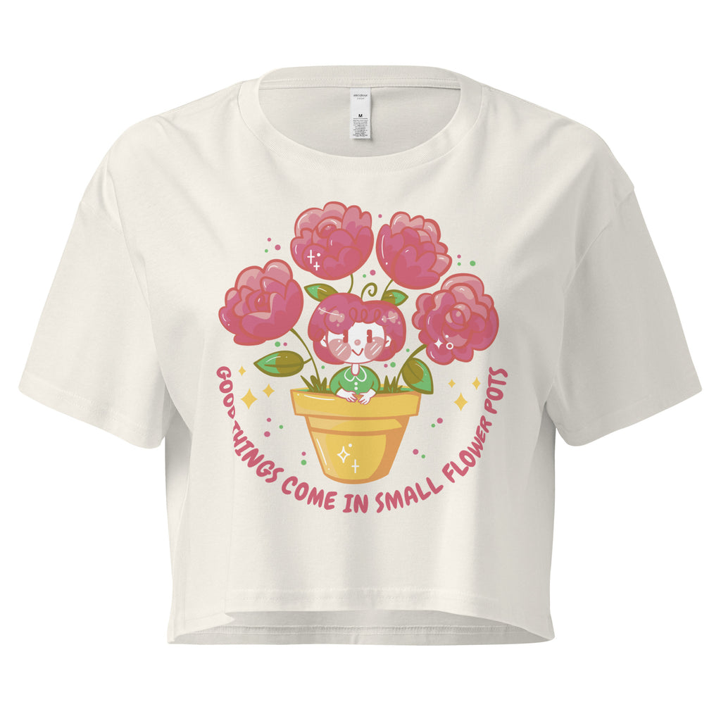 Ein ecru farbenes kurz geschnittenes Damen-T-Shirt (Crop Top) mit einer Illustration einer kleinen, lächelnden Figur in einem gelben Blumentopf. Um den Blumentopf herum sind mehrere große, rosa Blüten und grüne Blätter. Der Text "GOOD THINGS COME IN SMALL FLOWER POTS" ist in einem Bogen um das Motiv herum angeordnet, mit kleinen Sternen und Punkten.