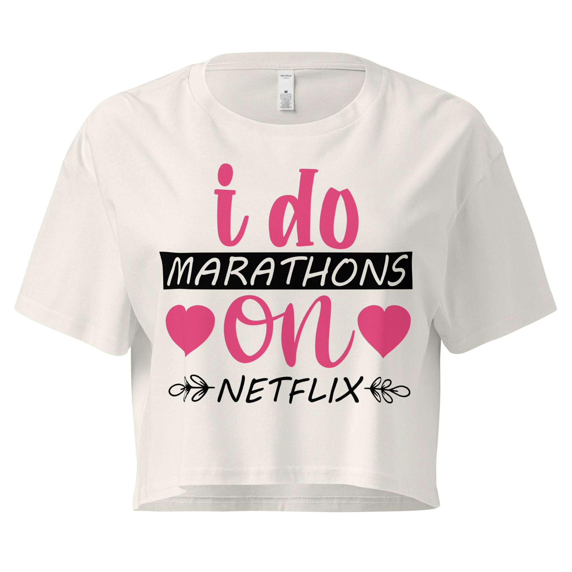 Ein ecru farbenes, kurz geschnittenes Damen-T-Shirt (Crop Top) mit dem Text "i do MARATHONS on NETFLIX" auf der Vorderseite. Der Text "i do" und "on" ist in Pink, "MARATHONS" in Schwarz auf einem schwarzen Balken, und "NETFLIX" ebenfalls in Schwarz, umgeben von kleinen schwarzen Herzen und floralen Ranken.