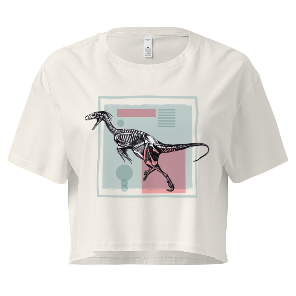 Ein ecru farbenes, kurz geschnittenes Damen-T-Shirt (Crop Top) mit einem weißen Kastenrahmen in der Mitte. Innerhalb des Rahmens befindet sich eine Illustration eines Dinosaurierskeletts, das auf einer Farbfeldfläche aus Rosa und Mintgrün platziert ist. Der Dinosaurier ist in einer Laufhaltung dargestellt.