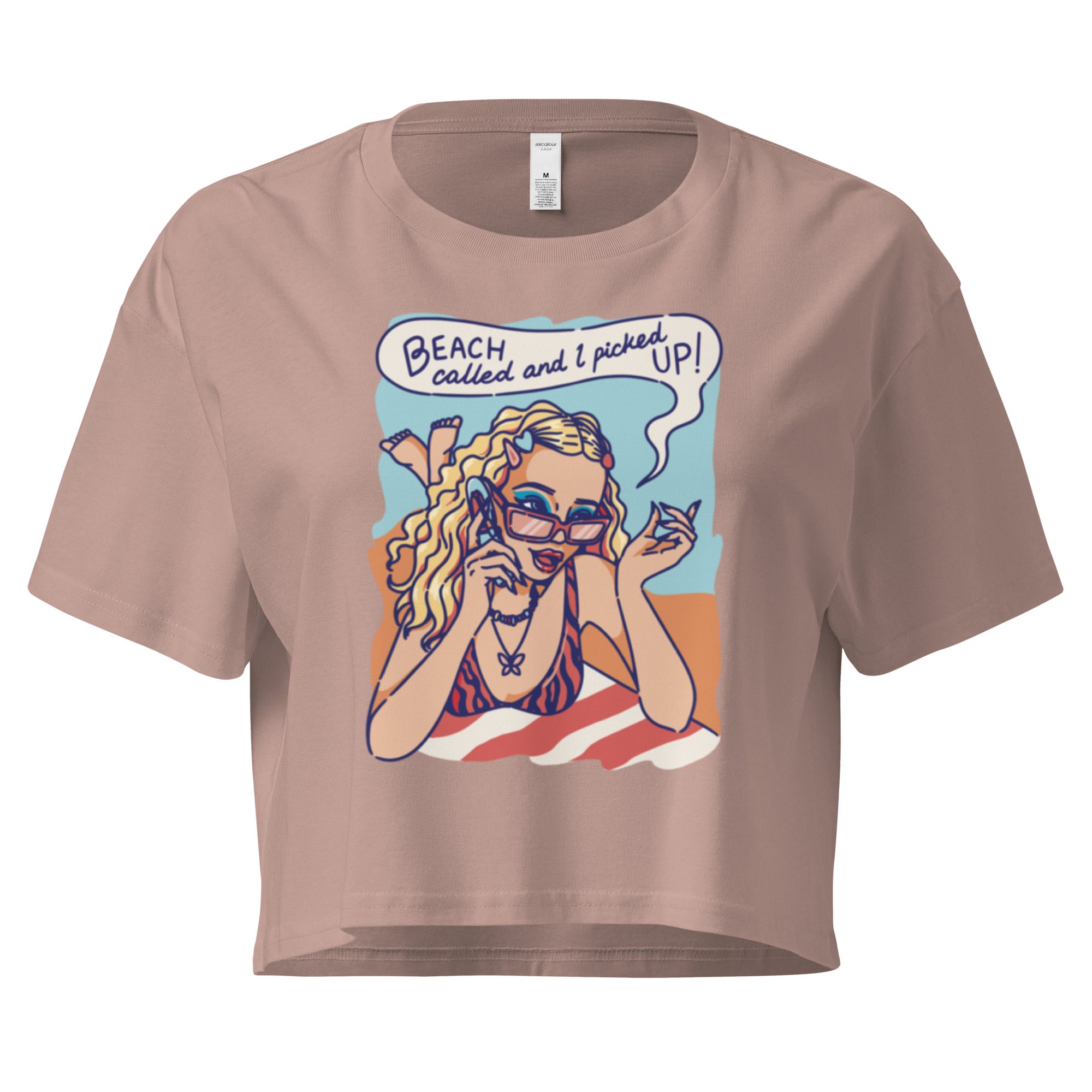 Ein hazy-pink farbenes kurz geschnittenes Damen-T-Shirt (Crop Top) mit einer Retro-Illustration einer blonden Frau mit Sonnenbrille und gestreiftem Bikini, die entspannt am Strand liegt und telefoniert. Über ihr ist eine Sprechblase mit dem Text "BEACH called and I picked UP!" zu sehen. Der Hintergrund zeigt Sand und Meer.