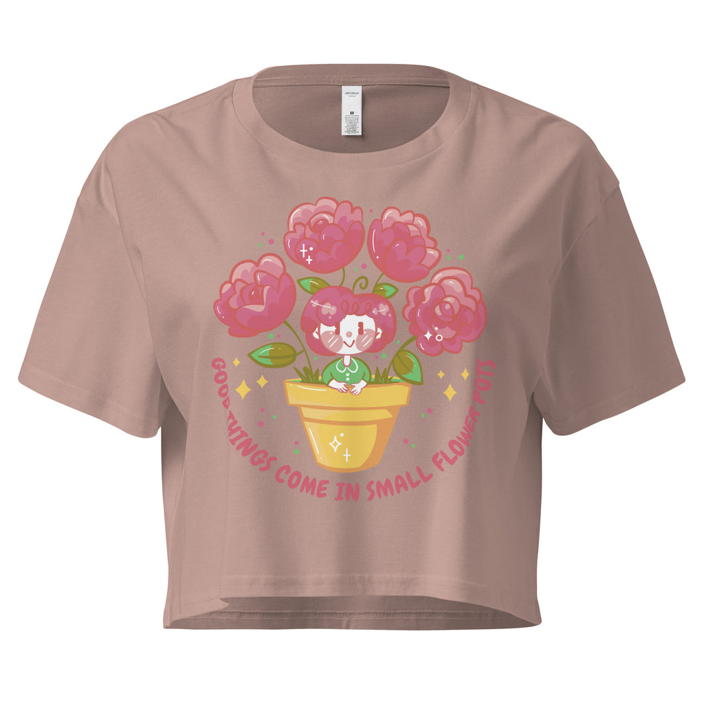Ein hazy-pink farbenes kurz geschnittenes Damen-T-Shirt (Crop Top) mit einer Illustration einer kleinen, lächelnden Figur in einem gelben Blumentopf. Um den Blumentopf herum sind mehrere große, rosa Blüten und grüne Blätter. Der Text "GOOD THINGS COME IN SMALL FLOWER POTS" ist in einem Bogen um das Motiv herum angeordnet, mit kleinen Sternen und Punkten.