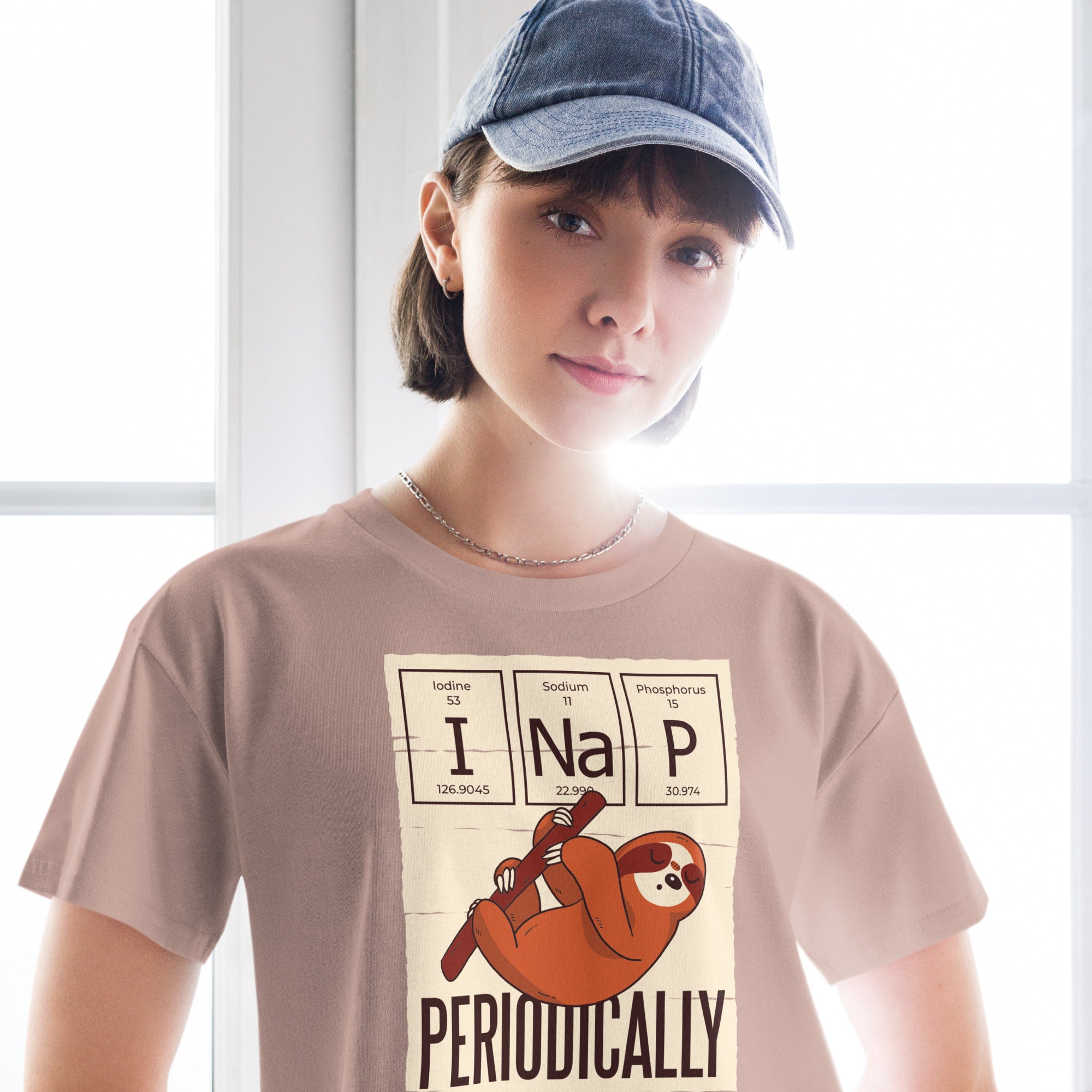 Ein hazy-pink farbenes kurz geschnittenes Damen-T-Shirt (Crop Top) mit einer Illustration eines Faultiers, das an einem Ast hängt. Darüber befinden sich drei Felder im Stil des Periodensystems der Elemente mit den Symbolen "I" (Iodine), "Na" (Sodium) und "P" (Phosphorus), die zusammen "I Na P" bilden. Unter dem Faultier steht das Wort "PERIODICALLY". Der Hintergrund des Motivs ist hellbeige.