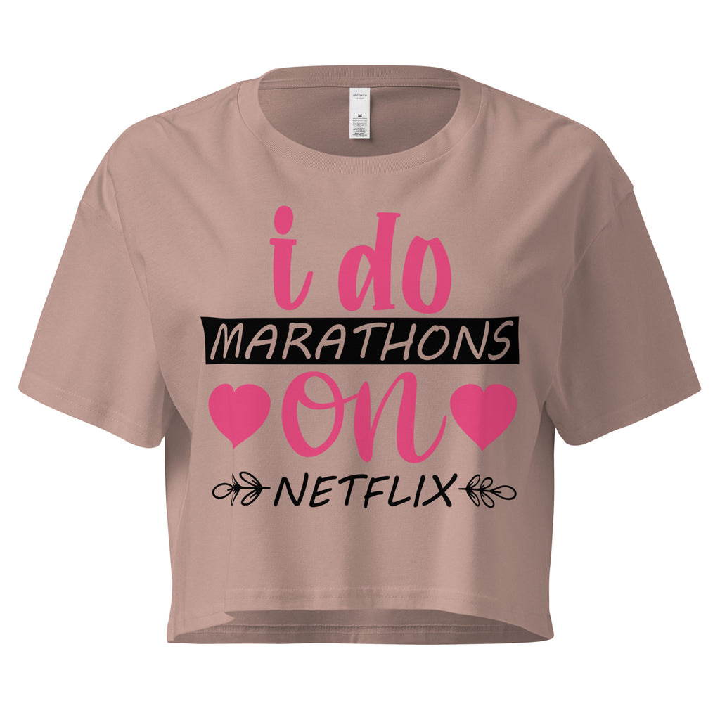 Ein hazy pink farbenes, kurz geschnittenes Damen-T-Shirt (Crop Top) mit dem Text "i do MARATHONS on NETFLIX" auf der Vorderseite. Der Text "i do" und "on" ist in Pink, "MARATHONS" in Schwarz auf einem schwarzen Balken, und "NETFLIX" ebenfalls in Schwarz, umgeben von kleinen schwarzen Herzen und floralen Ranken.