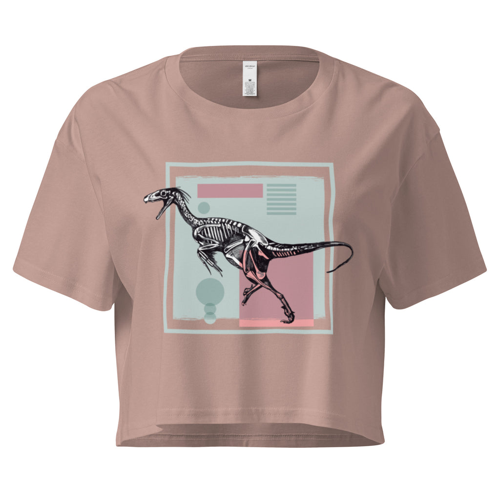 Ein hazy pink farbenes, kurz geschnittenes Damen-T-Shirt (Crop Top) mit einem weißen Kastenrahmen in der Mitte. Innerhalb des Rahmens befindet sich eine Illustration eines Dinosaurierskeletts, das auf einer Farbfeldfläche aus Rosa und Mintgrün platziert ist. Der Dinosaurier ist in einer Laufhaltung dargestellt.