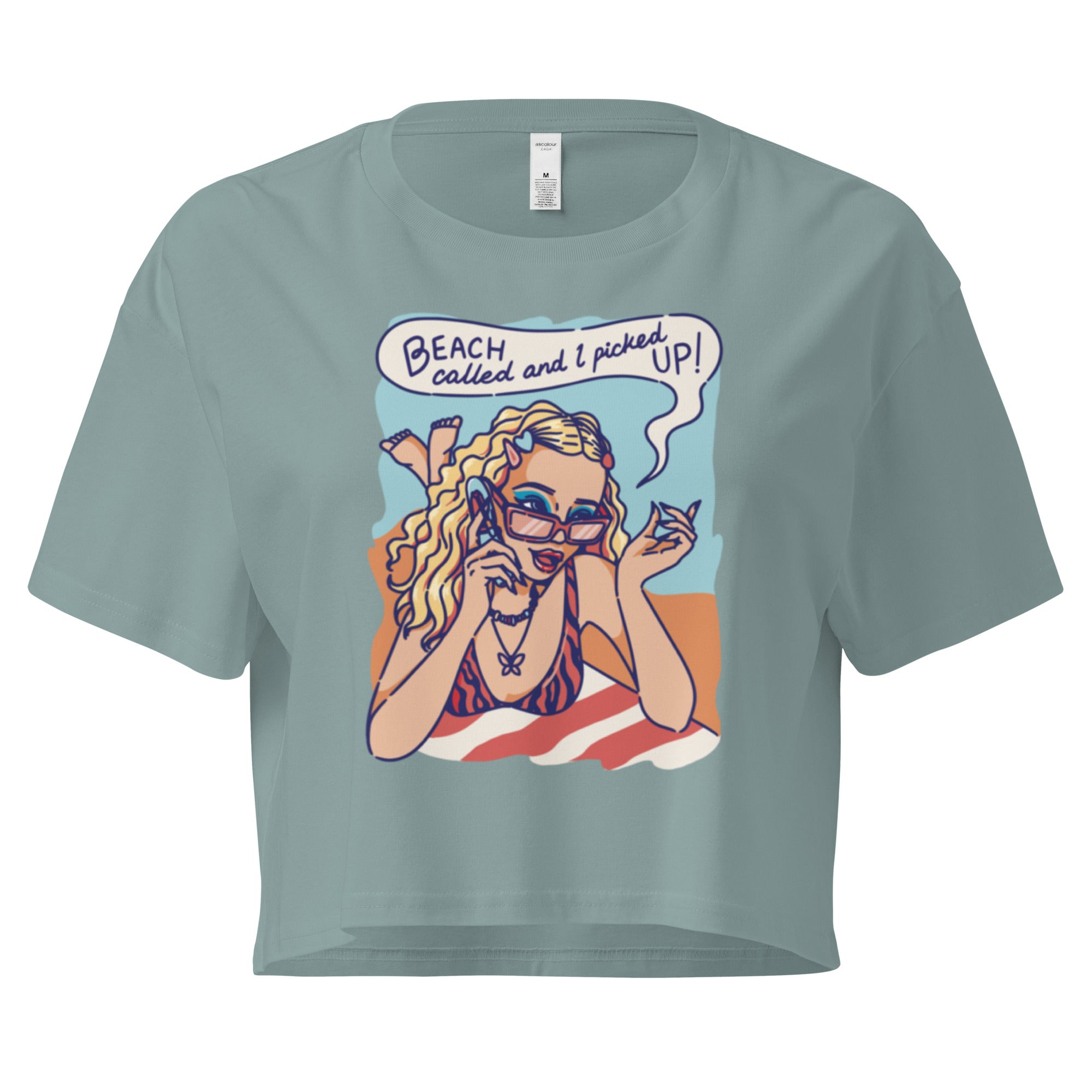 Ein mineral farbenes kurz geschnittenes Damen-T-Shirt (Crop Top) mit einer Retro-Illustration einer blonden Frau mit Sonnenbrille und gestreiftem Bikini, die entspannt am Strand liegt und telefoniert. Über ihr ist eine Sprechblase mit dem Text "BEACH called and I picked UP!" zu sehen. Der Hintergrund zeigt Sand und Meer.