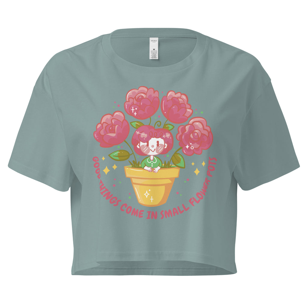 Ein mineral farbenes kurz geschnittenes Damen-T-Shirt (Crop Top) mit einer Illustration einer kleinen, lächelnden Figur in einem gelben Blumentopf. Um den Blumentopf herum sind mehrere große, rosa Blüten und grüne Blätter. Der Text "GOOD THINGS COME IN SMALL FLOWER POTS" ist in einem Bogen um das Motiv herum angeordnet, mit kleinen Sternen und Punkten.