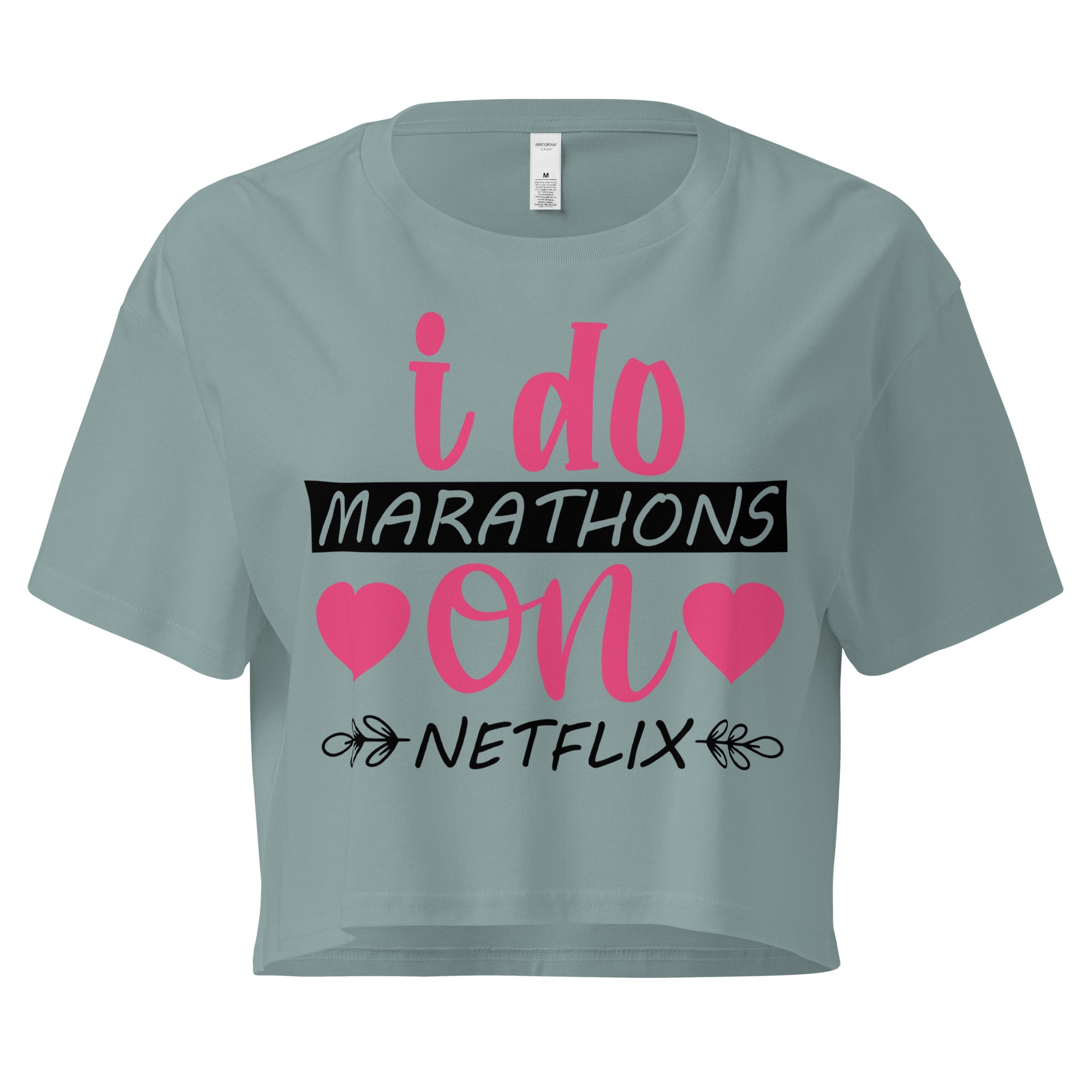 Ein mineralfarbenes, kurz geschnittenes Damen-T-Shirt (Crop Top) mit dem Text "i do MARATHONS on NETFLIX" auf der Vorderseite. Der Text "i do" und "on" ist in Pink, "MARATHONS" in Schwarz auf einem schwarzen Balken, und "NETFLIX" ebenfalls in Schwarz, umgeben von kleinen schwarzen Herzen und floralen Ranken.