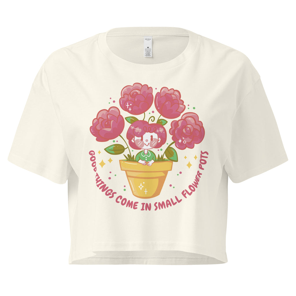 Ein natural farbenes kurz geschnittenes Damen-T-Shirt (Crop Top) mit einer Illustration einer kleinen, lächelnden Figur in einem gelben Blumentopf. Um den Blumentopf herum sind mehrere große, rosa Blüten und grüne Blätter. Der Text "GOOD THINGS COME IN SMALL FLOWER POTS" ist in einem Bogen um das Motiv herum angeordnet, mit kleinen Sternen und Punkten.
