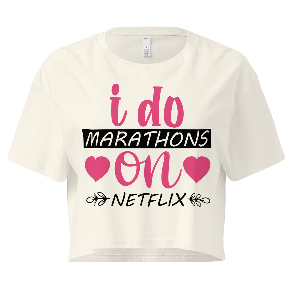 Ein natur farbenes, kurz geschnittenes Damen-T-Shirt (Crop Top) mit dem Text "i do MARATHONS on NETFLIX" auf der Vorderseite. Der Text "i do" und "on" ist in Pink, "MARATHONS" in Schwarz auf einem schwarzen Balken, und "NETFLIX" ebenfalls in Schwarz, umgeben von kleinen schwarzen Herzen und floralen Ranken.