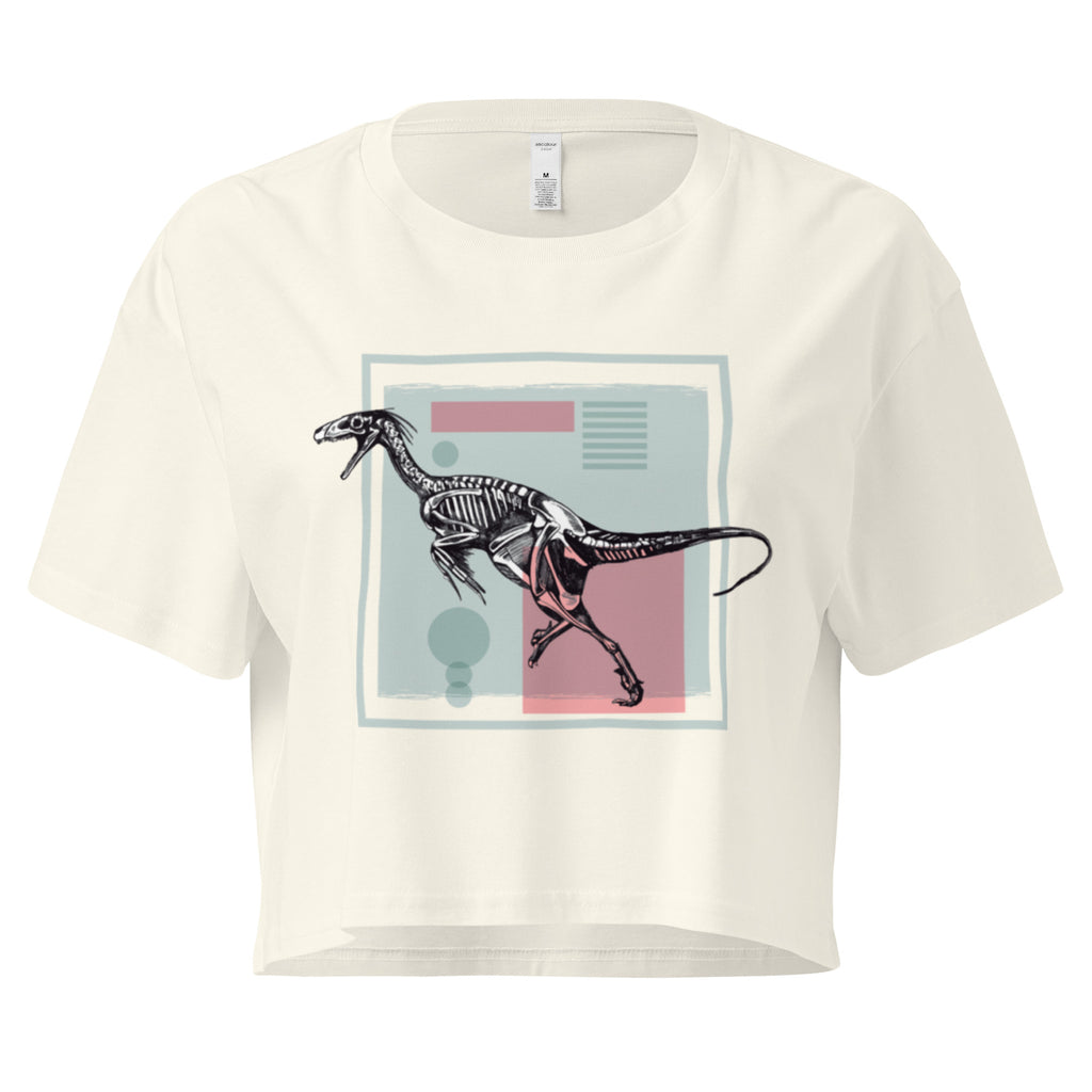 Ein natur farbenes, kurz geschnittenes Damen-T-Shirt (Crop Top) mit einem weißen Kastenrahmen in der Mitte. Innerhalb des Rahmens befindet sich eine Illustration eines Dinosaurierskeletts, das auf einer Farbfeldfläche aus Rosa und Mintgrün platziert ist. Der Dinosaurier ist in einer Laufhaltung dargestellt.