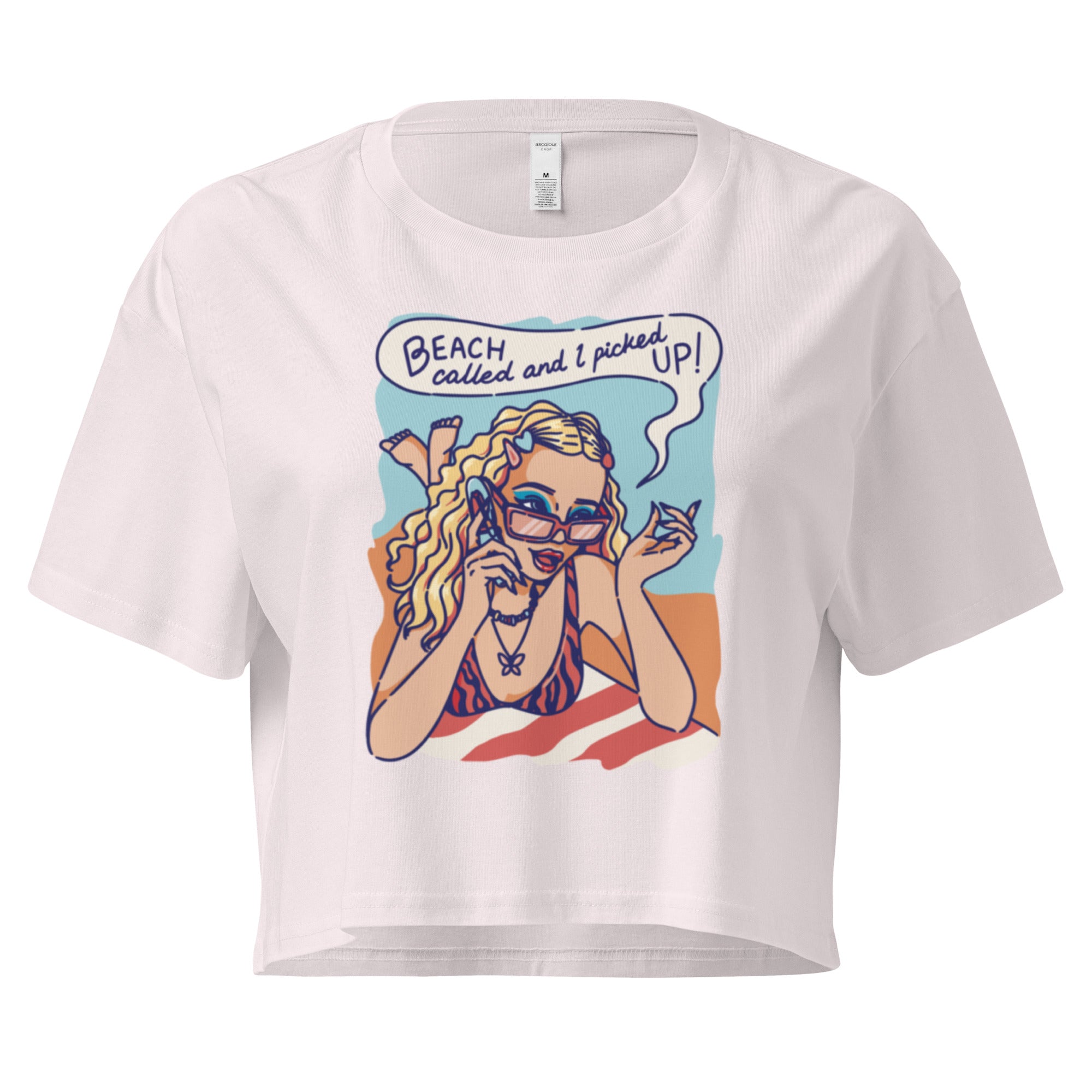 Ein orchid farbenes kurz geschnittenes Damen-T-Shirt (Crop Top) mit einer Retro-Illustration einer blonden Frau mit Sonnenbrille und gestreiftem Bikini, die entspannt am Strand liegt und telefoniert. Über ihr ist eine Sprechblase mit dem Text "BEACH called and I picked UP!" zu sehen. Der Hintergrund zeigt Sand und Meer.