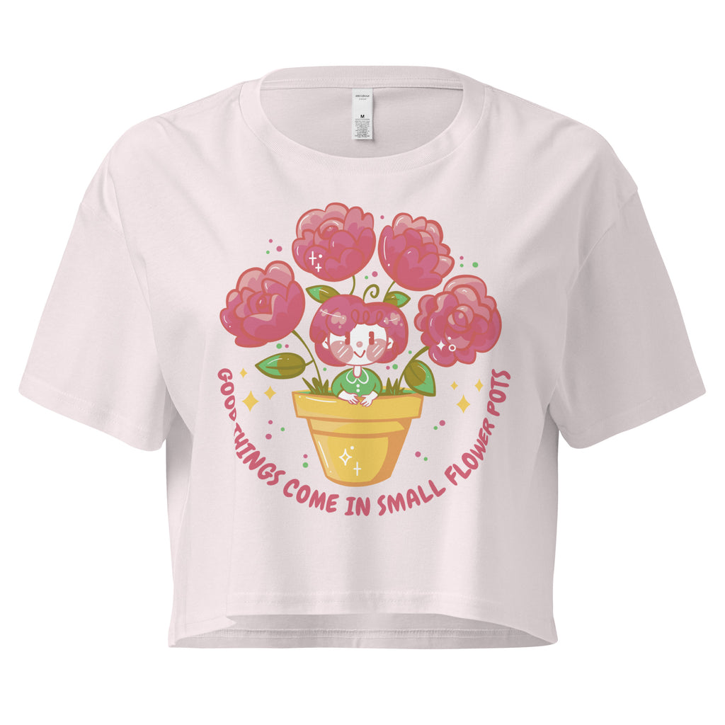 Ein orchid farbenes kurz geschnittenes Damen-T-Shirt (Crop Top) mit einer Illustration einer kleinen, lächelnden Figur in einem gelben Blumentopf. Um den Blumentopf herum sind mehrere große, rosa Blüten und grüne Blätter. Der Text "GOOD THINGS COME IN SMALL FLOWER POTS" ist in einem Bogen um das Motiv herum angeordnet, mit kleinen Sternen und Punkten.