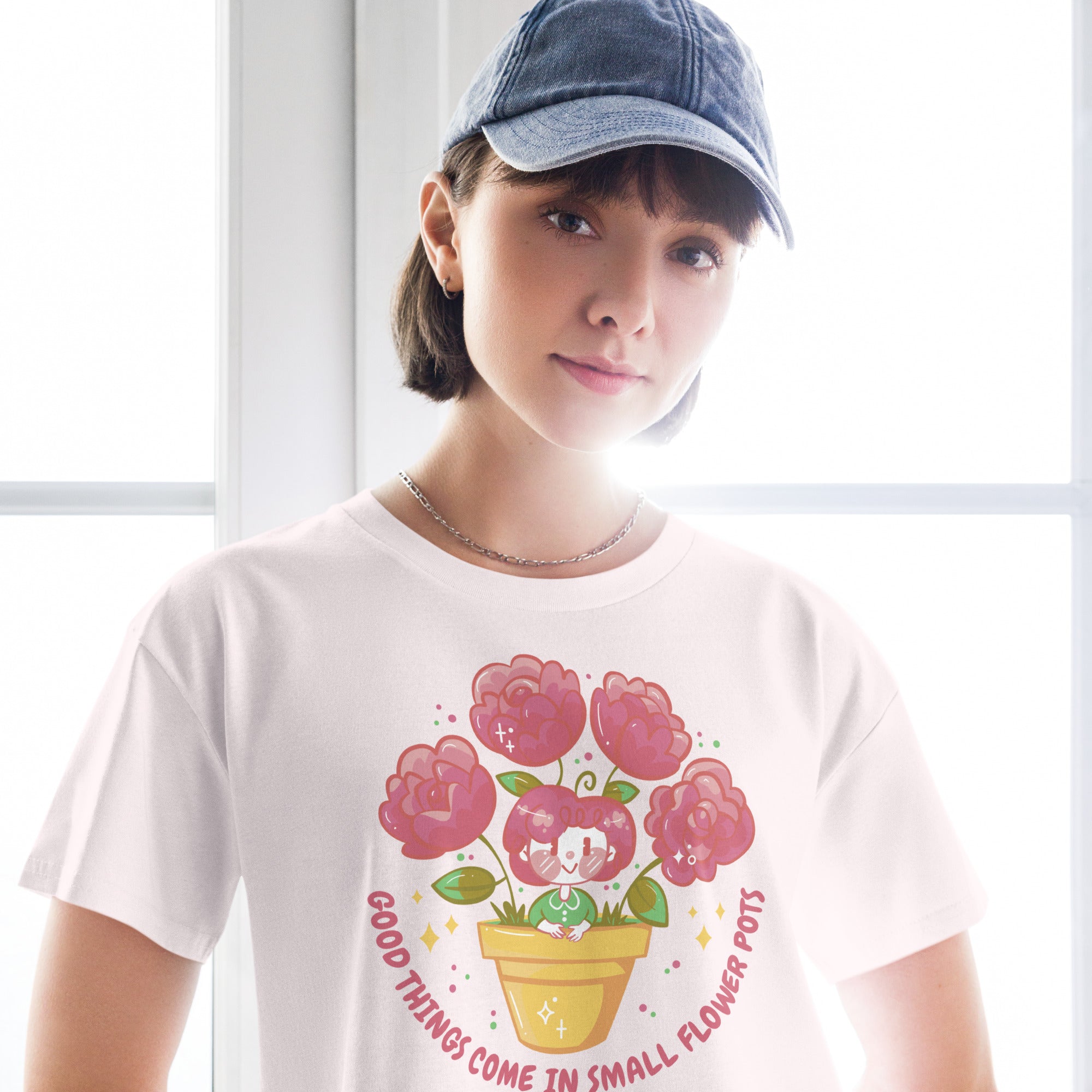 Ein orchid farbenes kurz geschnittenes Damen-T-Shirt (Crop Top) mit einer Illustration einer kleinen, lächelnden Figur in einem gelben Blumentopf. Um den Blumentopf herum sind mehrere große, rosa Blüten und grüne Blätter. Der Text "GOOD THINGS COME IN SMALL FLOWER POTS" ist in einem Bogen um das Motiv herum angeordnet, mit kleinen Sternen und Punkten.