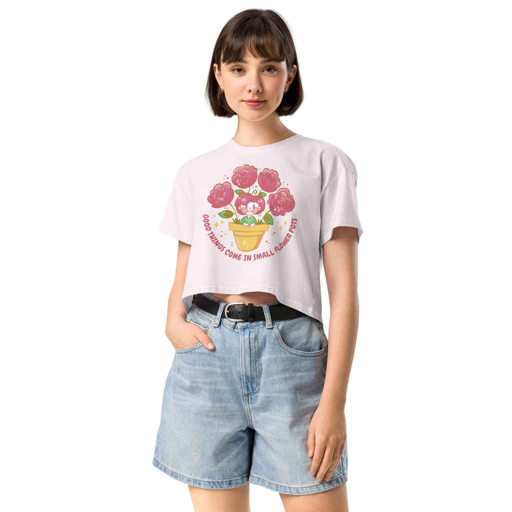 Ein orchid farbenes kurz geschnittenes Damen-T-Shirt (Crop Top) mit einer Illustration einer kleinen, lächelnden Figur in einem gelben Blumentopf. Um den Blumentopf herum sind mehrere große, rosa Blüten und grüne Blätter. Der Text "GOOD THINGS COME IN SMALL FLOWER POTS" ist in einem Bogen um das Motiv herum angeordnet, mit kleinen Sternen und Punkten.