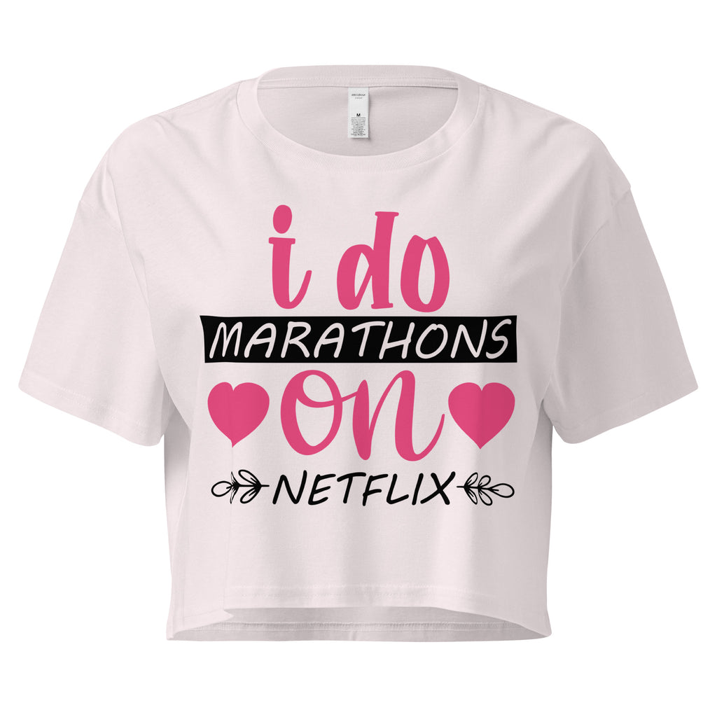 Ein orchid farbenes, kurz geschnittenes Damen-T-Shirt (Crop Top) mit dem Text "i do MARATHONS on NETFLIX" auf der Vorderseite. Der Text "i do" und "on" ist in Pink, "MARATHONS" in Schwarz auf einem schwarzen Balken, und "NETFLIX" ebenfalls in Schwarz, umgeben von kleinen schwarzen Herzen und floralen Ranken.