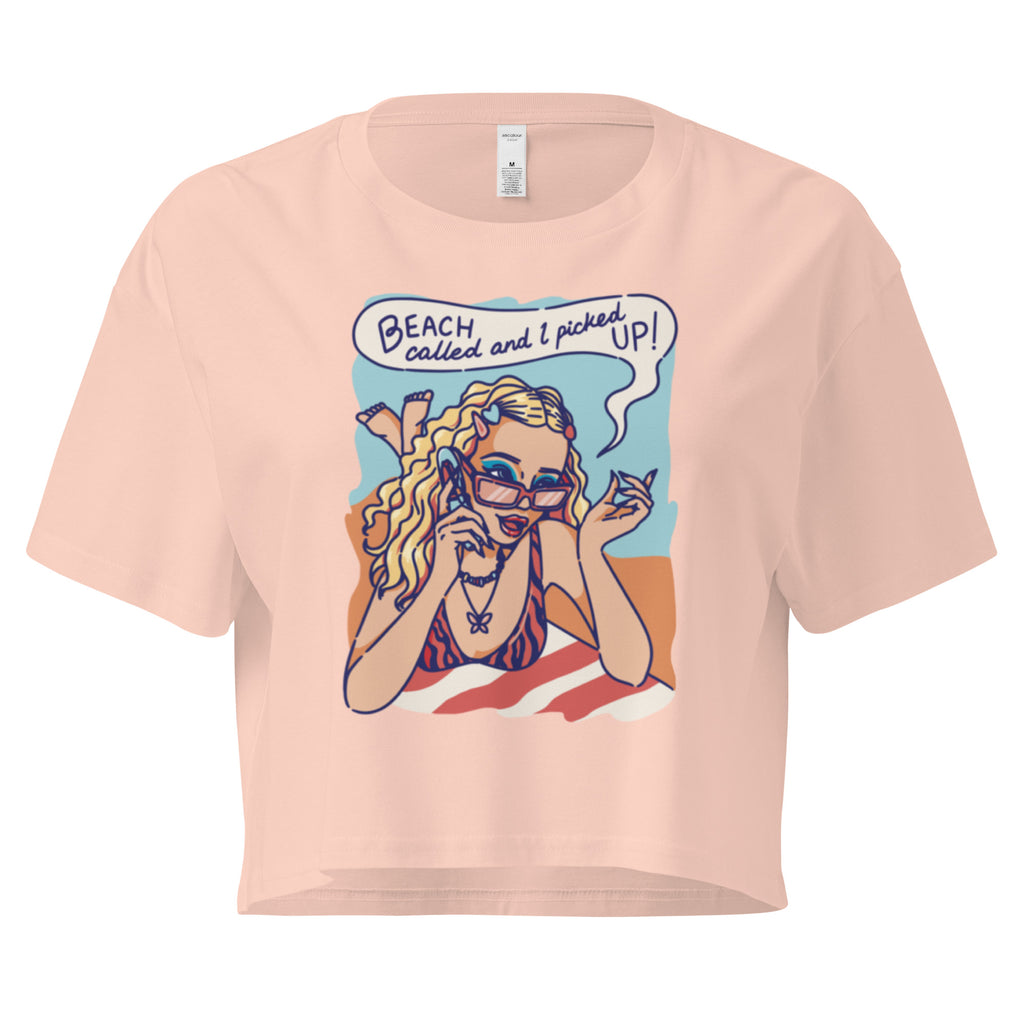 Ein pale-pink farbenes kurz geschnittenes Damen-T-Shirt (Crop Top) mit einer Retro-Illustration einer blonden Frau mit Sonnenbrille und gestreiftem Bikini, die entspannt am Strand liegt und telefoniert. Über ihr ist eine Sprechblase mit dem Text "BEACH called and I picked UP!" zu sehen. Der Hintergrund zeigt Sand und Meer.