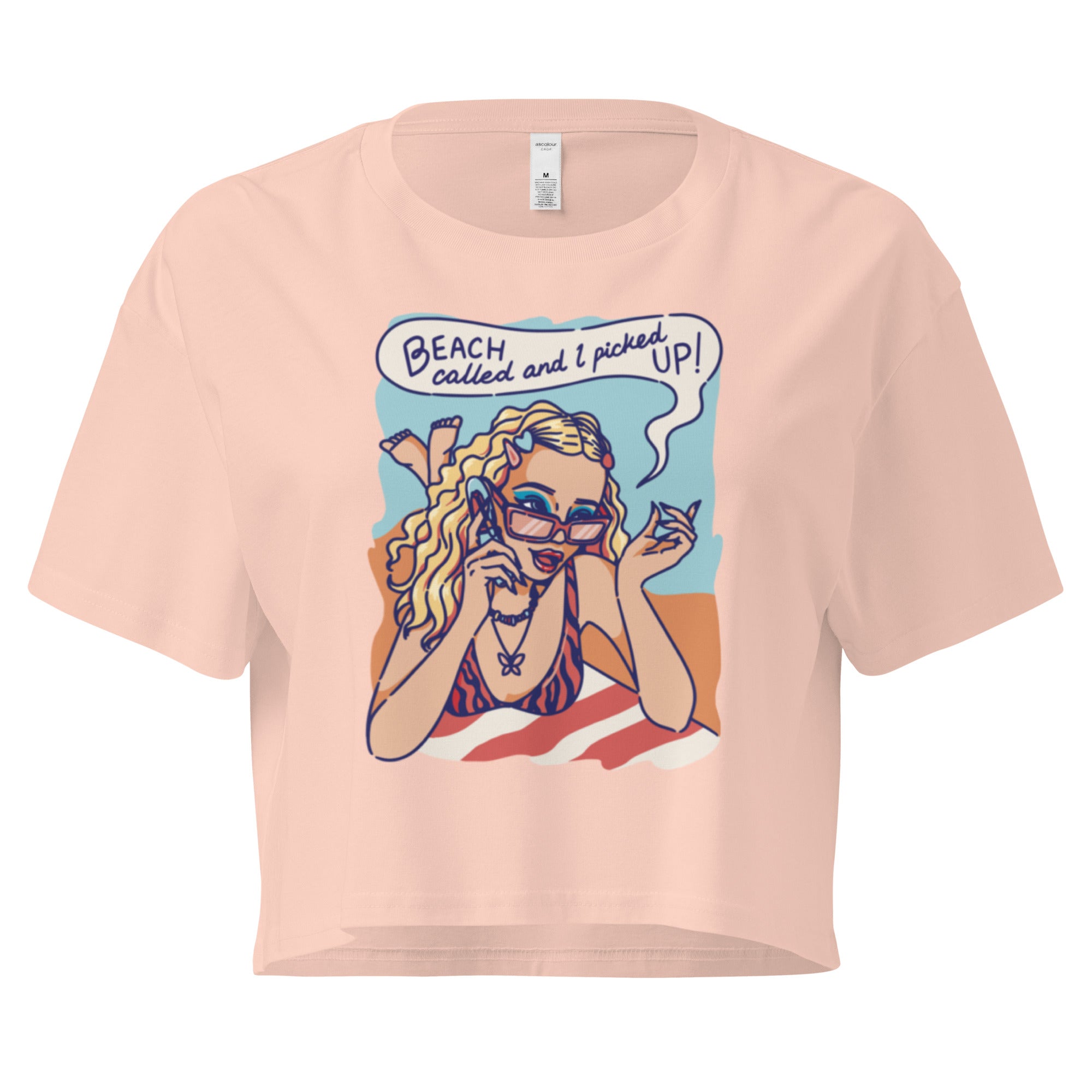 Ein pale-pink farbenes kurz geschnittenes Damen-T-Shirt (Crop Top) mit einer Retro-Illustration einer blonden Frau mit Sonnenbrille und gestreiftem Bikini, die entspannt am Strand liegt und telefoniert. Über ihr ist eine Sprechblase mit dem Text "BEACH called and I picked UP!" zu sehen. Der Hintergrund zeigt Sand und Meer.