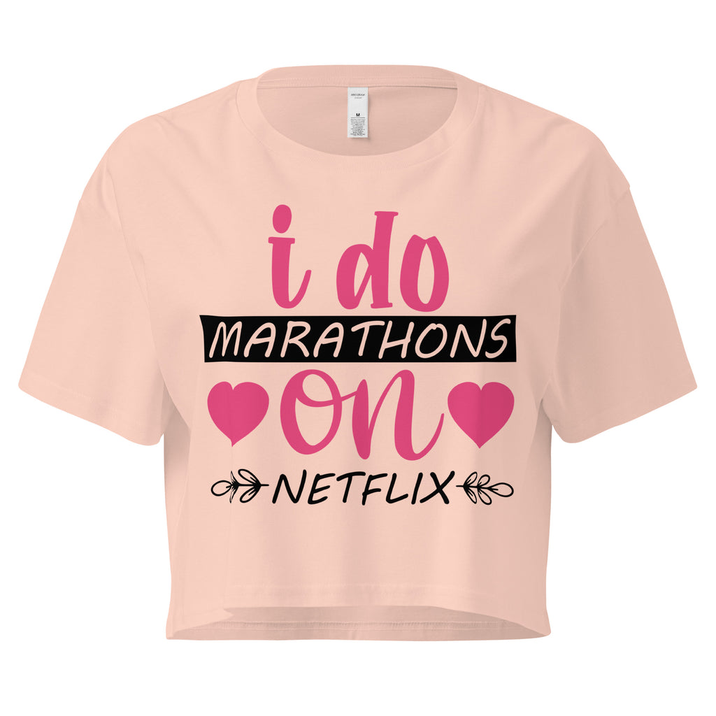 Ein pale pink farbenes, kurz geschnittenes Damen-T-Shirt (Crop Top) mit dem Text "i do MARATHONS on NETFLIX" auf der Vorderseite. Der Text "i do" und "on" ist in Pink, "MARATHONS" in Schwarz auf einem schwarzen Balken, und "NETFLIX" ebenfalls in Schwarz, umgeben von kleinen schwarzen Herzen und floralen Ranken.