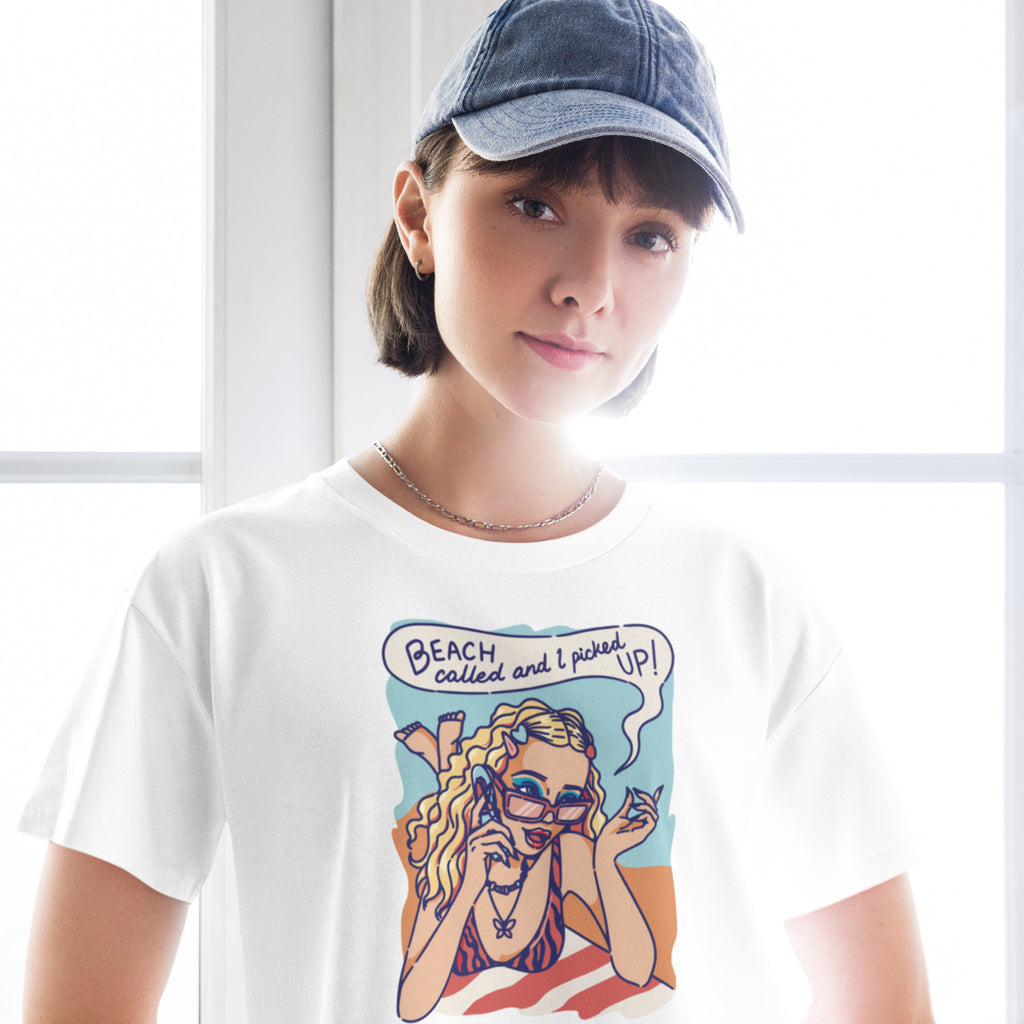 Ein weißes kurz geschnittenes Damen-T-Shirt (Crop Top) mit einer Retro-Illustration einer blonden Frau mit Sonnenbrille und gestreiftem Bikini, die entspannt am Strand liegt und telefoniert. Über ihr ist eine Sprechblase mit dem Text "BEACH called and I picked UP!" zu sehen. Der Hintergrund zeigt Sand und Meer.