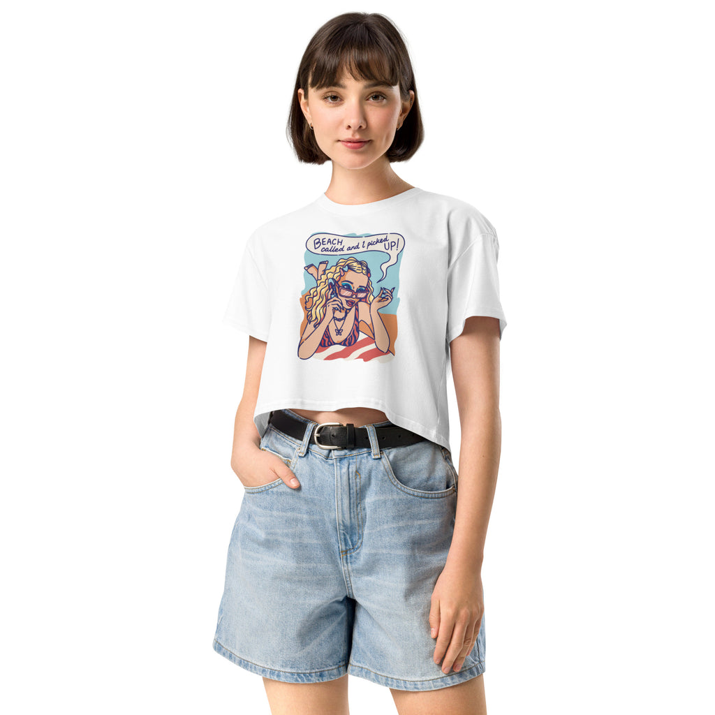 Ein weißes kurz geschnittenes Damen-T-Shirt (Crop Top) mit einer Retro-Illustration einer blonden Frau mit Sonnenbrille und gestreiftem Bikini, die entspannt am Strand liegt und telefoniert. Über ihr ist eine Sprechblase mit dem Text "BEACH called and I picked UP!" zu sehen. Der Hintergrund zeigt Sand und Meer.
