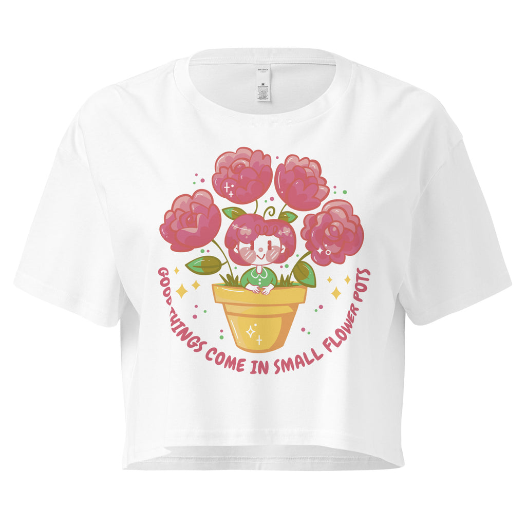 Ein weißes kurz geschnittenes Damen-T-Shirt (Crop Top) mit einer Illustration einer kleinen, lächelnden Figur in einem gelben Blumentopf. Um den Blumentopf herum sind mehrere große, rosa Blüten und grüne Blätter. Der Text "GOOD THINGS COME IN SMALL FLOWER POTS" ist in einem Bogen um das Motiv herum angeordnet, mit kleinen Sternen und Punkten.