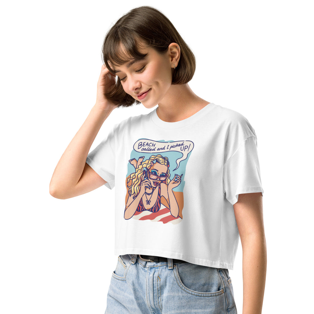 Ein weißes kurz geschnittenes Damen-T-Shirt (Crop Top) mit einer Retro-Illustration einer blonden Frau mit Sonnenbrille und gestreiftem Bikini, die entspannt am Strand liegt und telefoniert. Über ihr ist eine Sprechblase mit dem Text "BEACH called and I picked UP!" zu sehen. Der Hintergrund zeigt Sand und Meer.