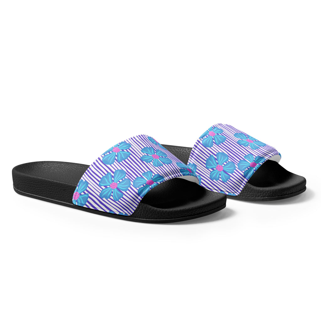 Ein Paar schwarze Damen-Badeschlappen mit einem breiten Riemen. Der Riemen ist mit einem Muster aus vertikalen lila und weißen Streifen versehen, die von großen, stilisierten blauen Blüten mit pinkfarbenen Zentren überlagert werden. Die Sohle ist schwarz und hat eine leicht texturierte Oberfläche.