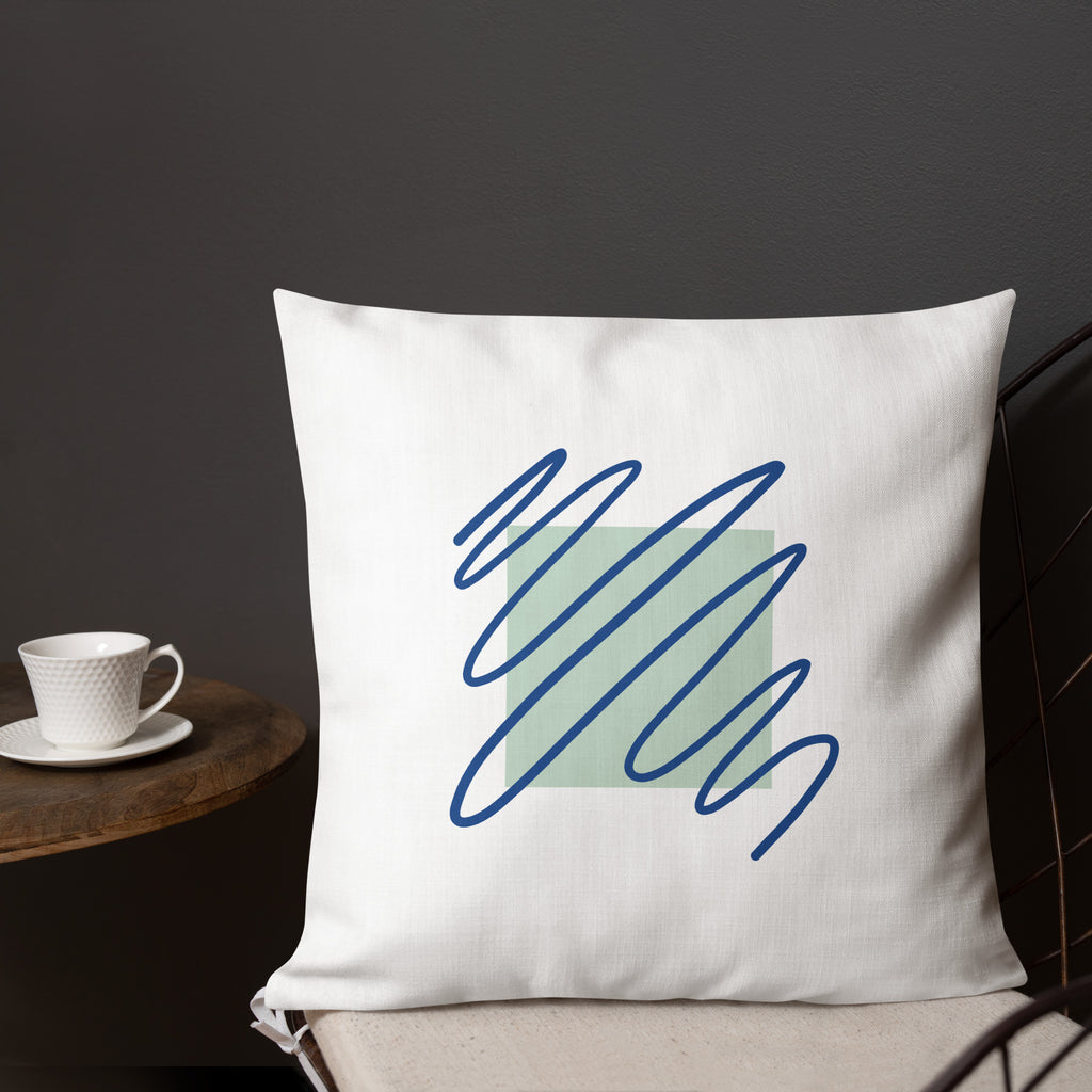 Ein weißes Kissen mit einem quadratischen, hellgrünen Muster im Zentrum. Darüber sind blaue, gekritzelte Linien zu sehen. Das Kissen liegt auf einem Stuhl, und links im Bild ist ein Teil eines kleinen Tisches mit einer weißen Tasse zu erkennen. Das Kissen ist in der Größe "18 x 18" erhältlich. Designed, by Just a Girl