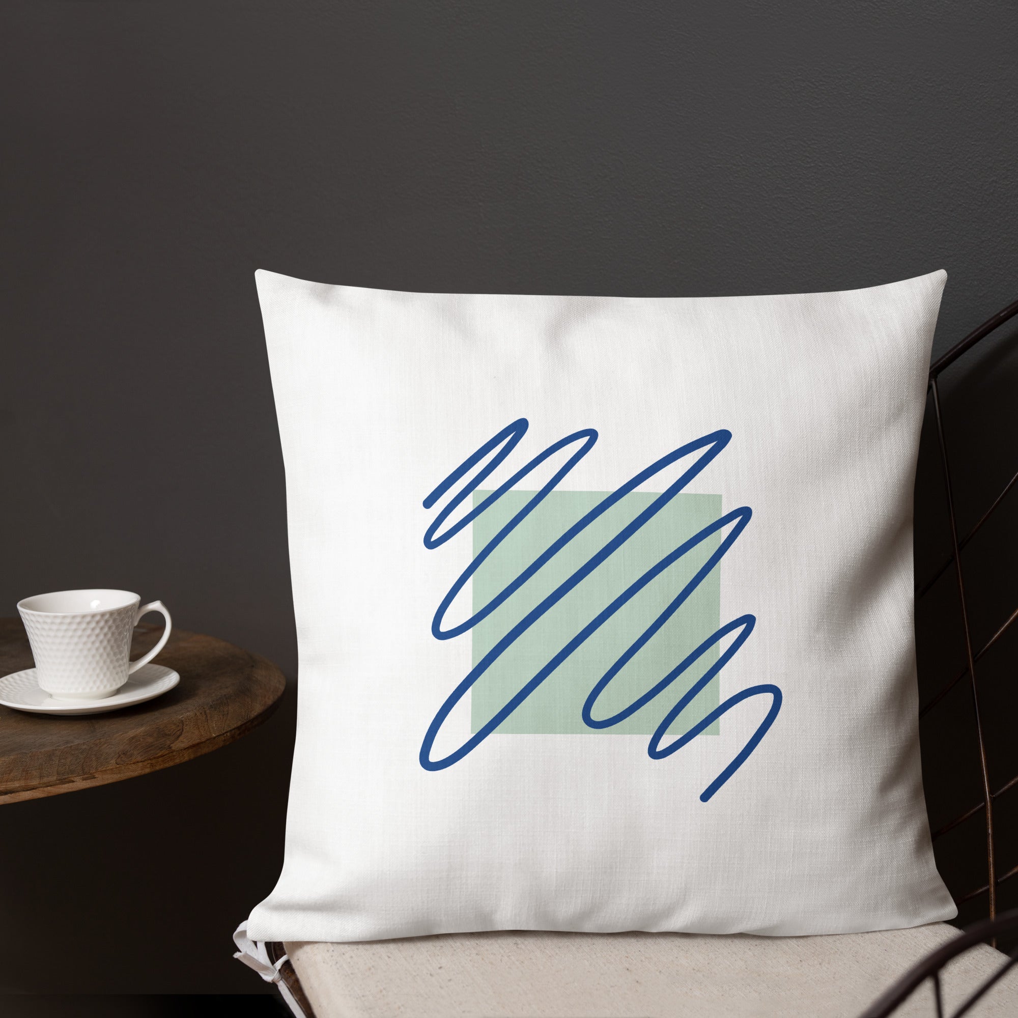 Ein weißes Kissen mit einem quadratischen, hellgrünen Muster im Zentrum. Darüber sind blaue, gekritzelte Linien zu sehen. Das Kissen liegt auf einem Stuhl, und links im Bild ist ein Teil eines kleinen Tisches mit einer weißen Tasse zu erkennen. Das Kissen ist in der Größe "18 x 18" erhältlich. Designed, by Just a Girl