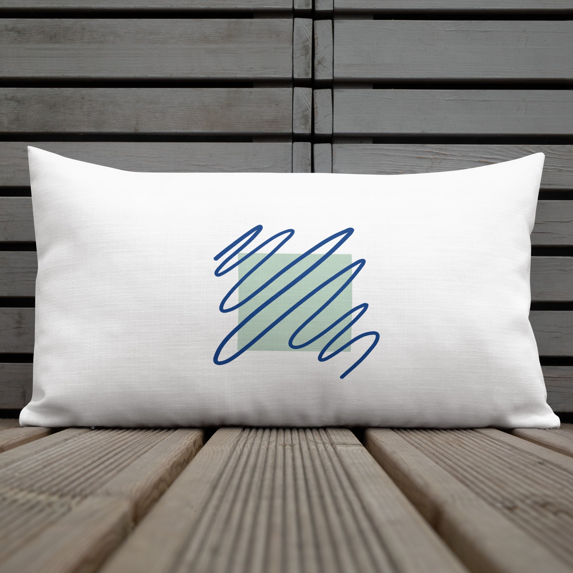 Ein weißes Kissen mit einem quadratischen, hellgrünen Muster im Zentrum. Darüber sind blaue, gekritzelte Linien zu sehen. Das Kissen liegt auf einem Stuhl, und links im Bild ist ein Teil eines kleinen Tisches mit einer weißen Tasse zu erkennen. Das Kissen ist in der Größe "20 x 12" erhältlich. Designed, by Just a Girl