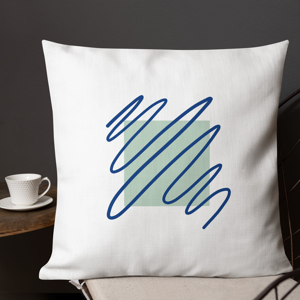 Ein weißes Kissen mit einem quadratischen, hellgrünen Muster im Zentrum. Darüber sind blaue, gekritzelte Linien zu sehen. Das Kissen liegt auf einem Stuhl, und links im Bild ist ein Teil eines kleinen Tisches mit einer weißen Tasse zu erkennen. Das Kissen ist in der Größe "22 x 22" erhältlich. Designed, by Just a Girl