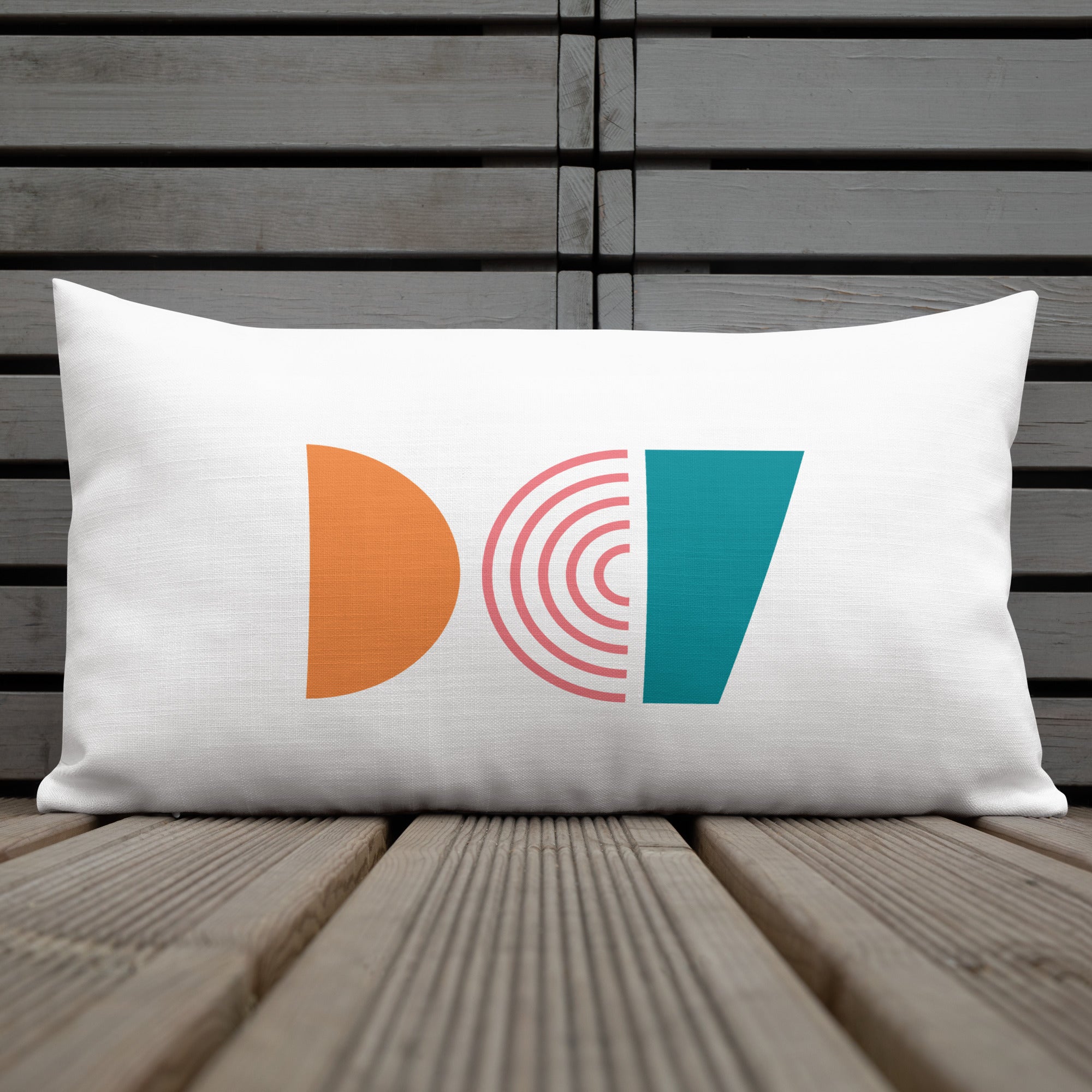 Ein dekoratives Kissen mit einem geometrischen Muster, das aus drei verschiedenen Formen besteht. Oben befindet sich eine orangefarbene Halbkugel, in der Mitte ein Regenbogenmuster in Rosatönen und unten ein blaugrünes Trapez. Das Kissen liegt auf einem Stuhl, und ein Teil eines Tischs mit einer weißen Tasse ist auf der linken Seite des Bildes zu sehen. Dieses Kissen ist in der Größe 20" x 12" erhältlich. Designed, by Just a Girl.