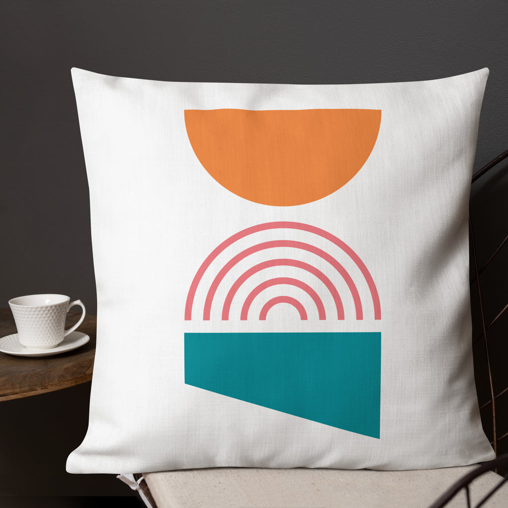 Ein dekoratives Kissen mit einem geometrischen Muster, das aus drei verschiedenen Formen besteht. Oben befindet sich eine orangefarbene Halbkugel, in der Mitte ein Regenbogenmuster in Rosatönen und unten ein blaugrünes Trapez. Das Kissen liegt auf einem Stuhl, und ein Teil eines Tischs mit einer weißen Tasse ist auf der linken Seite des Bildes zu sehen. Dieses Kissen ist in der Größe 22" x 22" erhältlich. Designed, by Just a Girl.