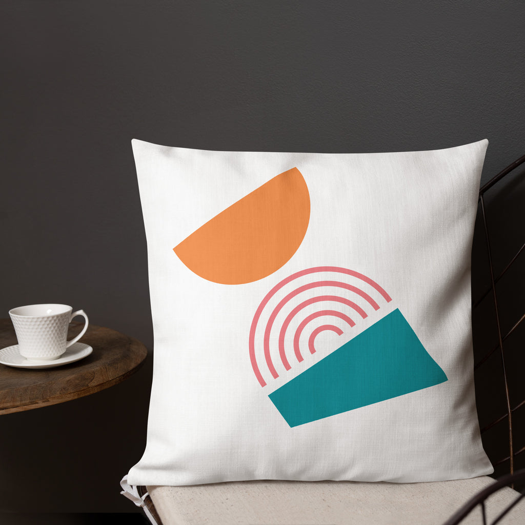 Ein dekoratives Kissen mit einem geometrischen Muster, das aus drei verschiedenen Formen besteht. Oben befindet sich eine orangefarbene Halbkugel, in der Mitte ein Regenbogenmuster in Rosatönen und unten ein blaugrünes Trapez. Das Kissen liegt auf einem Stuhl, und ein Teil eines Tischs mit einer weißen Tasse ist auf der linken Seite des Bildes zu sehen. Dieses Kissen ist in der Größe 18" x 18" erhältlich. Designed, by Just a Girl.
